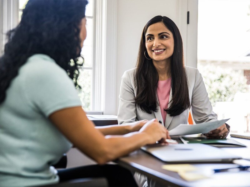 Two professional women engage in a conversation while sitting together in a modern open office space