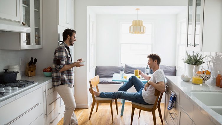 2 male talking to each other at home(one sitting on chair with cup of coffee and other is standing with a bowl of food)