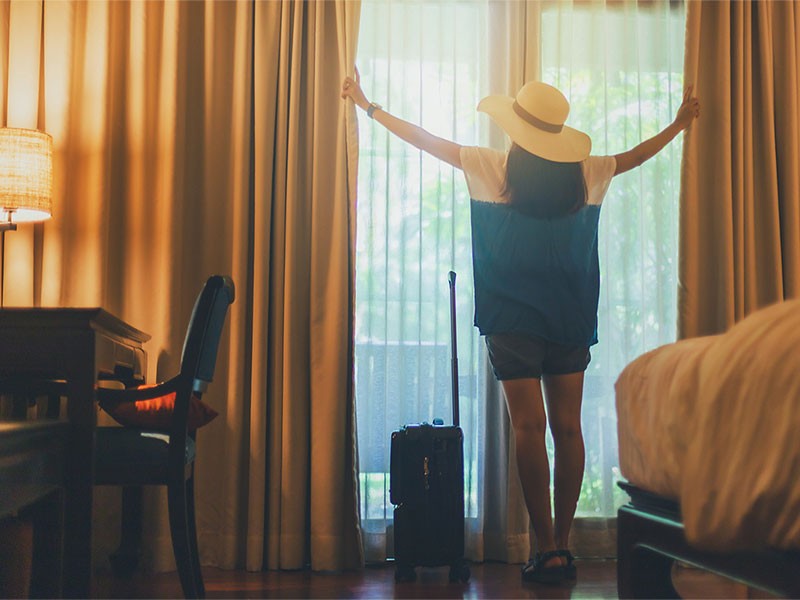 A lady looking outside while opening the curtains of a window from hotel room